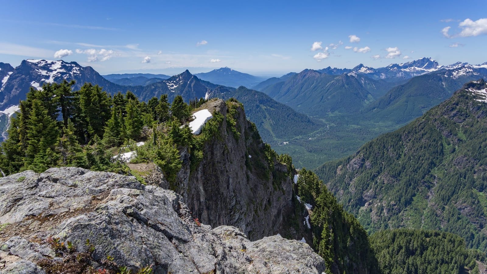 Looking over a rocky outcrop near the top of Mount Dickerman to forested valleys and snow-patched peaks