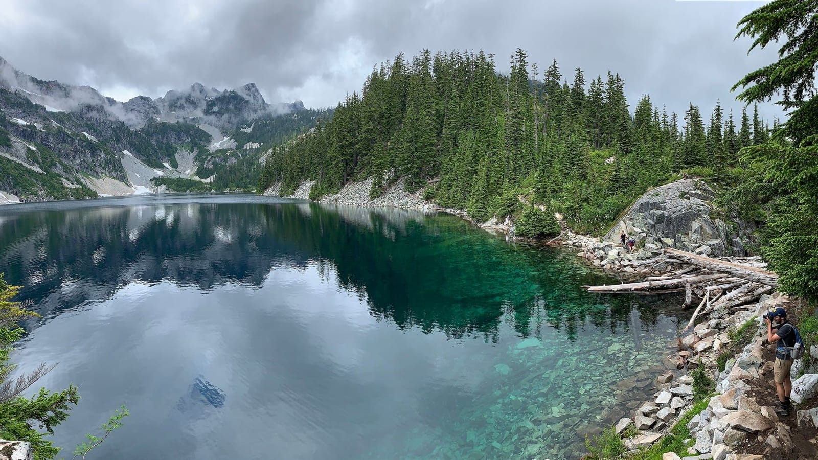 Clear green water of Snow Lake below forested slopes and cloud-capped peaks near Snoqualmie Pass